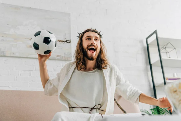 Happy Jesus in crown of thorns sitting on sofa with soccer ball and watching football match at home — Stock Photo