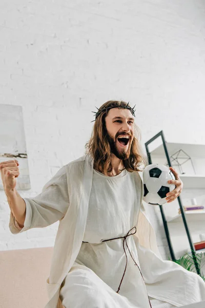 Взволнованный Иисус в терновом венце жестом руками и смотреть футбольный матч дома — стоковое фото