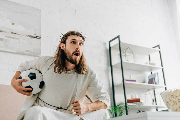 Jesús emocional en corona de espinas sentado en el sofá con pelota de fútbol y viendo el partido de fútbol en casa - foto de stock