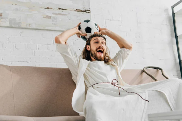 Aufgeregter Jesus in Dornenkrone hält Fußballball über dem Kopf und sieht Fußballspiel zu Hause — Stockfoto