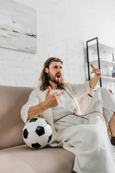 Agresivo Jesús en corona de espinas viendo fútbol y mostrando dedos medios partido en casa - foto de stock