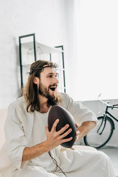 Seitenansicht des glücklichen Jesus in Dornenkrone, der Ball hält und Rugby auf dem Sofa zu Hause guckt — Stockfoto