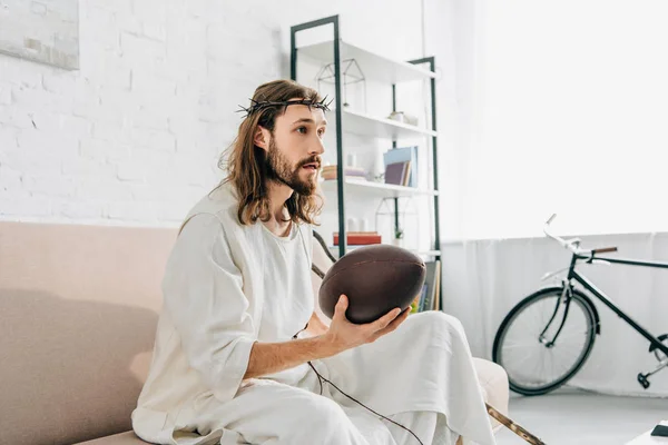 Selective focus of Jesus in crown of thorns holding ball and watching rugby on sofa at home — Stock Photo