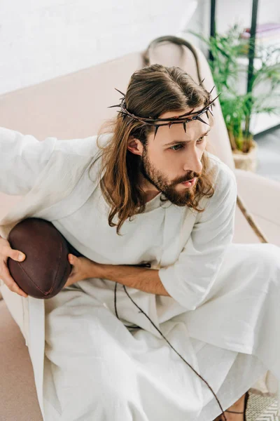 Vue grand angle de Jésus concentré en couronne d'épines tenant le ballon et regardant le football américain sur le canapé à la maison — Photo de stock