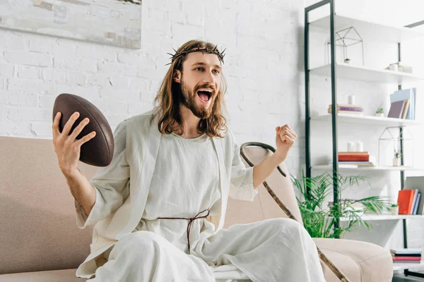Excitado Jesús en corona de espinas haciendo gestos a mano y viendo fútbol americano en el sofá en casa - foto de stock