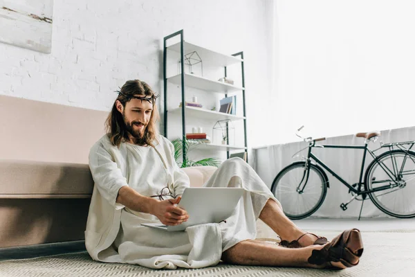 Smiling Jesus sitting on floor and using laptop near sofa at home — Stock Photo