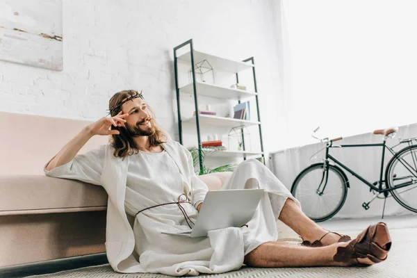 Verträumter Jesus sitzt auf dem Boden und benutzt Laptop in der Nähe von Sofa zu Hause — Stockfoto