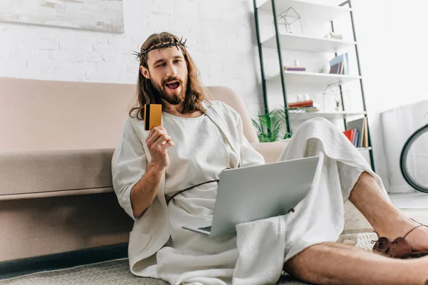 Happy Jesus showing credit card, winking and doing online shopping on laptop near sofa at home — Stock Photo