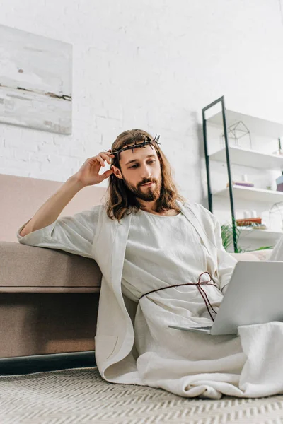 Confident Jesus in crown of thorns sitting on floor and using laptop near sofa at home — Stock Photo