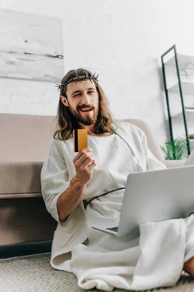 Fröhlicher Jesus zeigt Kreditkarte, zwinkert und erledigt Online-Einkäufe auf Laptop in der Nähe des heimischen Sofas — Stockfoto
