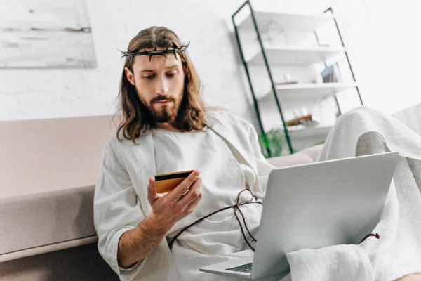 Thoughtful Jesus looking at credit card and doing online shopping on laptop near sofa at home — Stock Photo