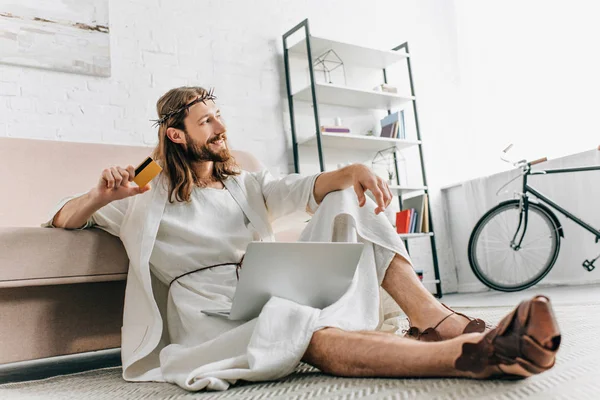 Улыбаясь Иисус сидит на полу с кредитной картой и делает покупки в Интернете на ноутбуке рядом с диваном дома — стоковое фото