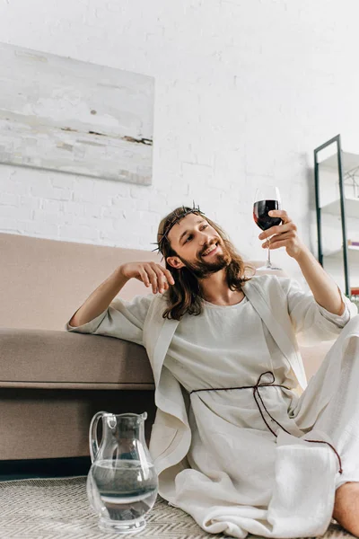 Glücklicher Jesus in Dornenkronen und Robe auf dem Boden sitzend in der Nähe von Wasserkrug und zu Hause Wein aus dem Glas trinkend — Stockfoto