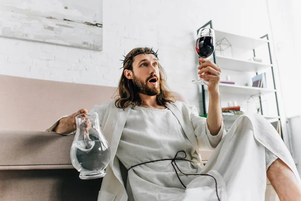 Shocked Jesus in crowns of thorns and robe holding jug of water and looking at wine glass in hand at home — Stock Photo