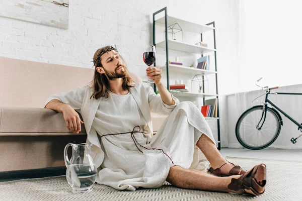 Jesús escéptico en coronas de espinas y túnica sentado en el suelo cerca de la jarra de agua y beber vino de vidrio en casa - foto de stock