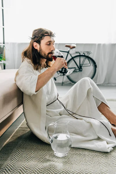 Seitenansicht des Jesus in Dornenkronen und Robe auf dem Boden sitzend in der Nähe von Wasserkrug und zu Hause Wein aus Glas trinkend — Stockfoto