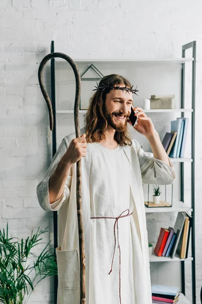 Smiling Jesus in robe with wooden staff talking on smartphone at home — Stock Photo