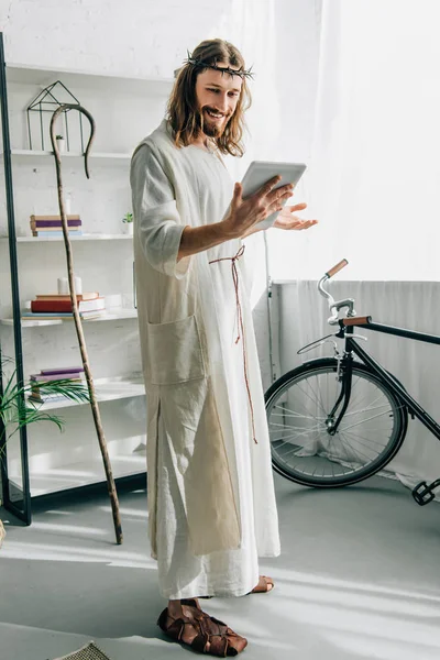 Счастливый Иисус в терновом венце и халате с помощью цифровой скрижали и жестом вручную дома — стоковое фото