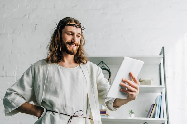 Smiling Jesus in crown of thorns and robe using digital tablet at home — Stock Photo
