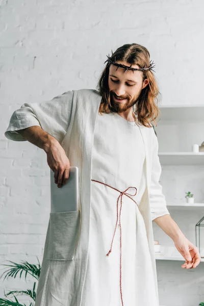 Selective focus of happy Jesus in crown of thorns putting digital tablet in pocket of robe at home — Stock Photo