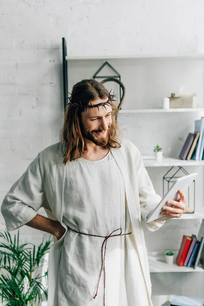 Jésus joyeux en couronne d'épines et robe à l'aide d'une tablette numérique à la maison — Photo de stock