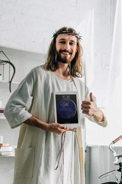 Jesús sonriente en corona de espinas haciendo gesto de pulgar y mostrando la tableta ipad en casa - foto de stock