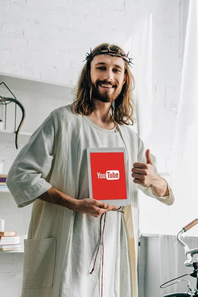 Cheerful Jesus in crown of thorns doing thumb gesture and showing digital tablet with youtube at home — Stock Photo