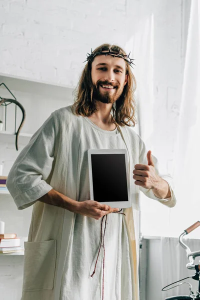 Feliz Jesús en corona de espinas haciendo gesto de pulgar y mostrando tableta digital con pantalla en blanco en casa - foto de stock