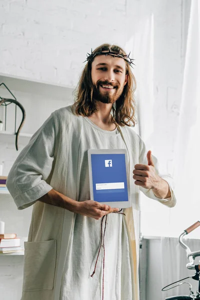 Smiling Jesus in crown of thorns doing thumb gesture and showing digital tablet with facebook website at home — Stock Photo