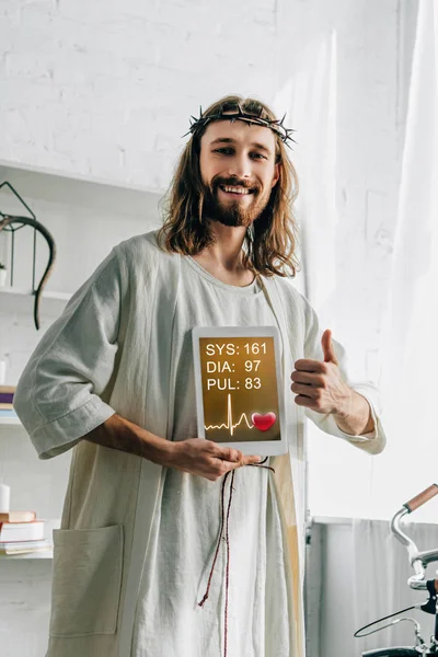 Fröhlicher Jesus in Dornenkrone macht Daumengeste und zeigt digitales Tablet mit medizinischer Anwendung zu Hause — Stockfoto