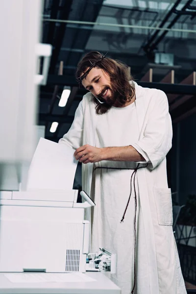 Fröhlicher Jesus in Dornenkrone und Robe im Gespräch mit Smartphone und Kopierer im modernen Büro — Stockfoto