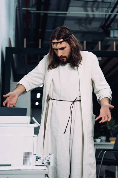 Emotional Jesus in crown of thorns and robe gesturing by hands and looking at copy machine in modern office — Stock Photo