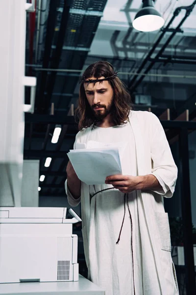 Focado Jesus na coroa de espinhos e robe verificando papéis perto de máquina de cópia no escritório moderno — Fotografia de Stock