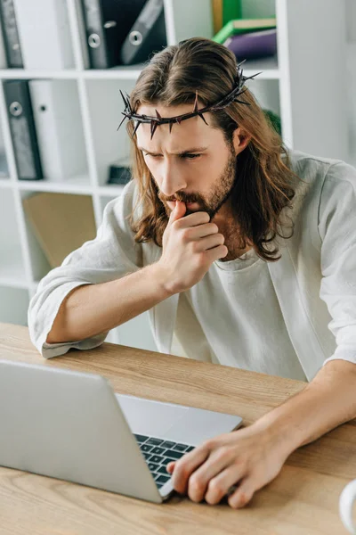 Thoughtful Jesus in crown of thorns using laptop at table in modern office — Stock Photo