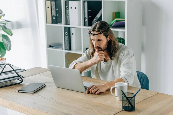 Задумчивый Иисус в терновом венце с помощью ноутбука за столом в современном офисе — стоковое фото