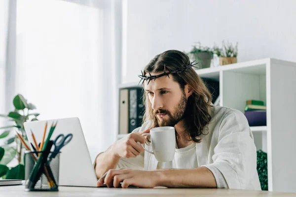 Confident Jesus in crown of thorns with coffee cup using laptop at table in modern office — Stock Photo