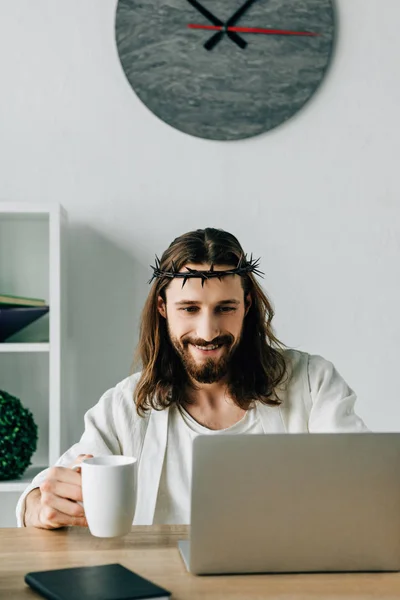 Cheerful Jesus in crown of thorns with coffee cup using laptop at table in modern office — Stock Photo