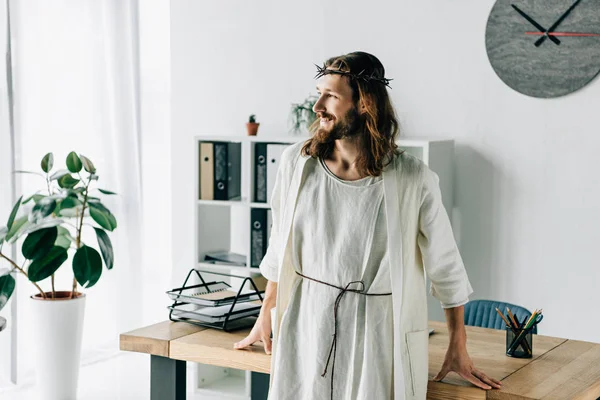 Enfoque selectivo de Jesús feliz en corona de espinas y túnica mirando hacia otro lado cerca de la mesa de trabajo en la oficina moderna - foto de stock