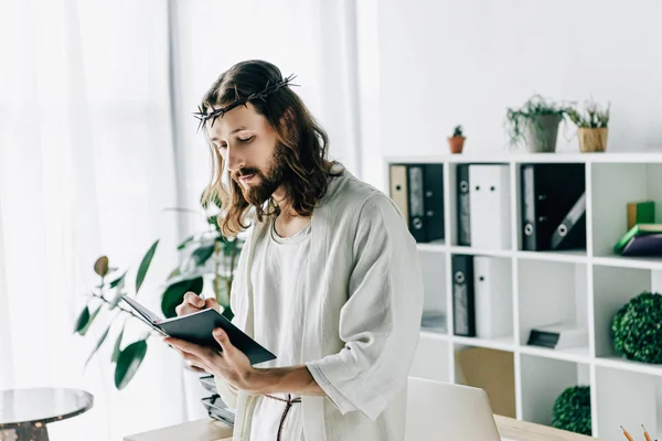 Serious Jesus in crown of thorns and robe writing in textbook near working table in modern office — Stock Photo