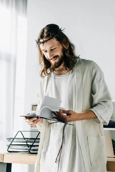 Enfoque selectivo de Jesús feliz en la corona de espinas y túnica lectura libro de texto cerca de mesa de trabajo en la oficina moderna - foto de stock