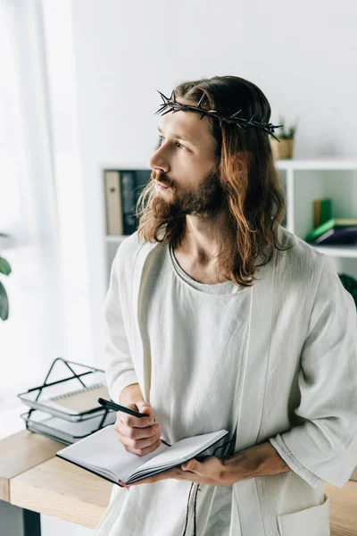 Pensive Jesus in crown of thorns and robe holding textbook and looking away in modern office — Stock Photo
