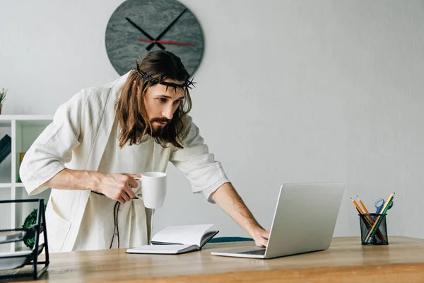 Focused Jesus in crown of thorns and robe holding coffee cup and using laptop at table in modern office — Stock Photo