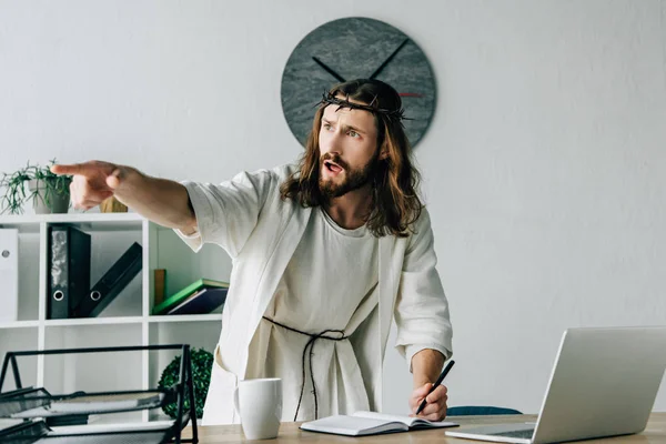 Агрессивный Иисус в терновом венце и мантии, пишущий в учебнике и указывающий пальцем за столом с ноутбуком в современном офисе — стоковое фото