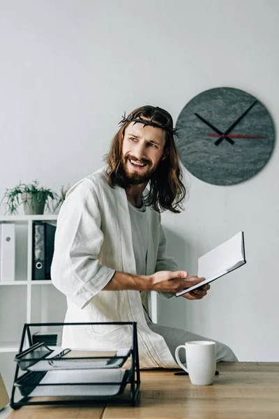 Angry Jesus in crown of thorns and robe sitting on table and pointing at textbook in modern office — Stock Photo