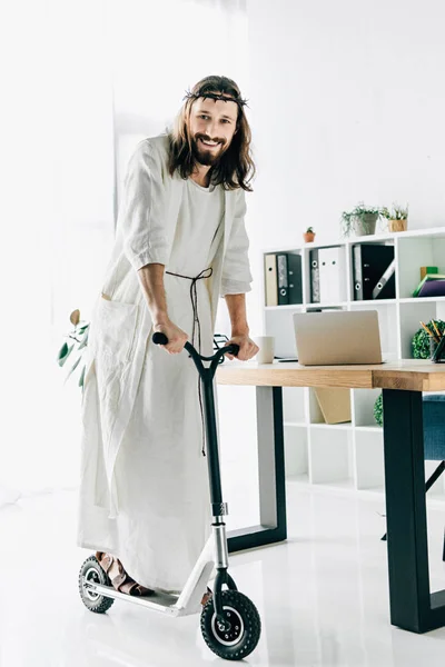 Feliz Jesus na coroa de espinhos e robe montando em scooter pontapé no escritório moderno — Fotografia de Stock