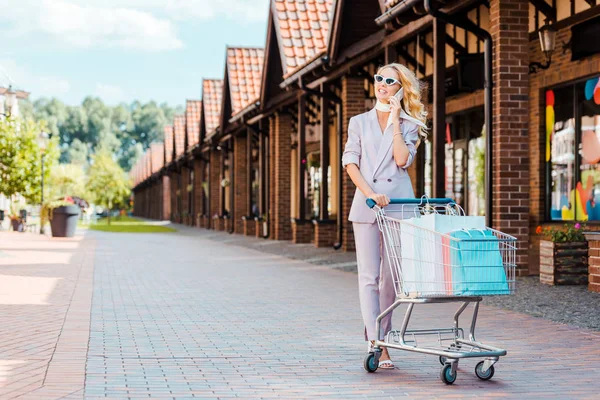 Belle jeune femme avec panier plein de sacs en papier parlant par téléphone sur la rue — Photo de stock