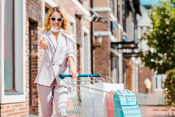 Bela jovem com carrinho de compras cheio de sacos de papel mostrando polegar para cima — Fotografia de Stock