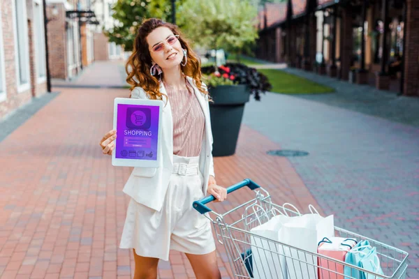 Hermosa joven con carrito de compras lleno de bolsas de papel que muestra la tableta con la aplicación de compras en la pantalla en la cámara - foto de stock