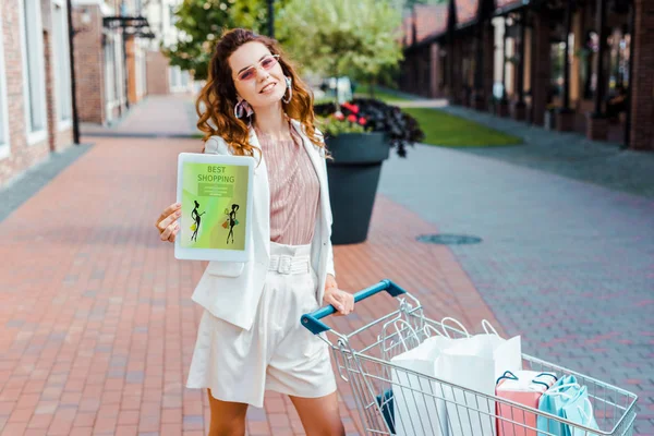 Bela jovem com carrinho de compras cheio de sacos de papel mostrando tablet com melhor aplicativo de compras na tela na câmera — Fotografia de Stock