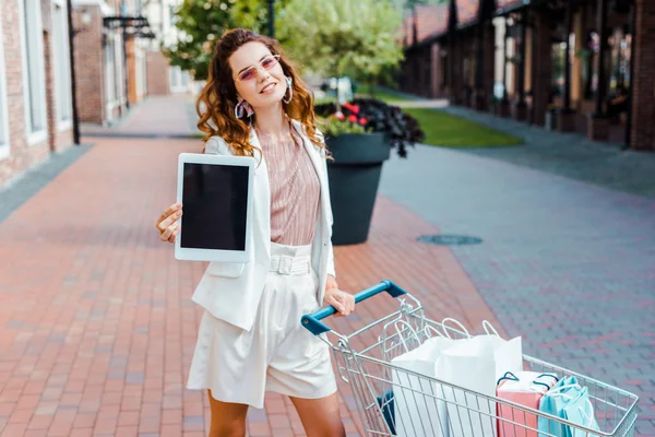 Belle jeune femme avec panier plein de sacs en papier montrant tablette avec écran blanc à la caméra — Photo de stock